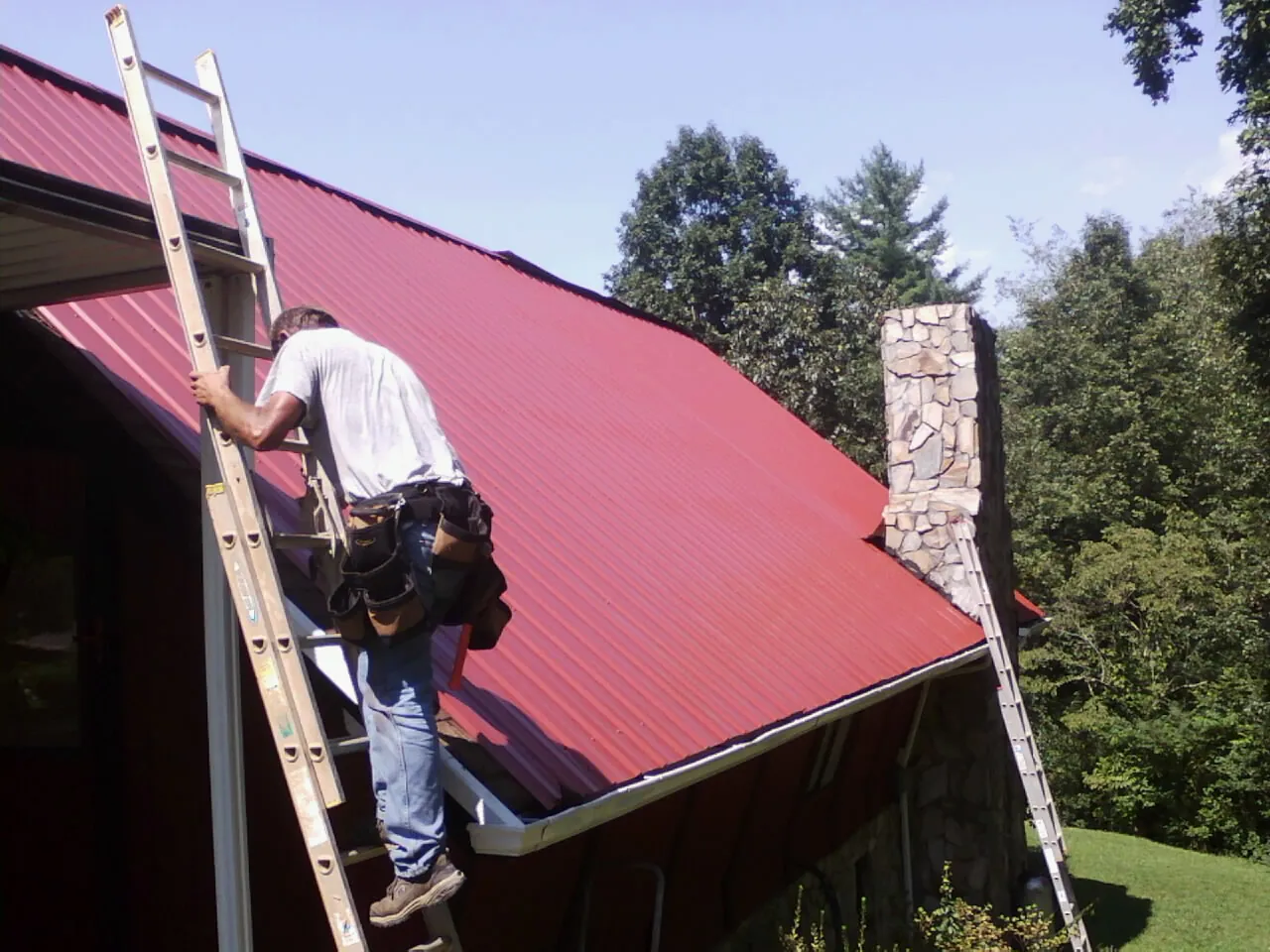 Roofer on a ladder beside a red metal roof and stone chimney — Soffit & Fascia Repair by Roofing Brothers in Mears